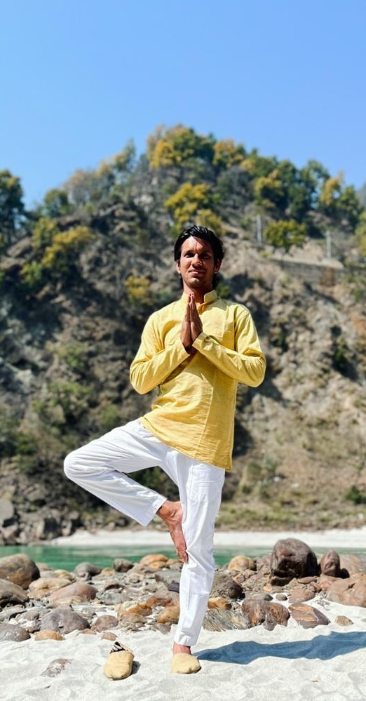 Devesh practising a steady standing yoga posture on river stones beside the Ganges and Himalayan foothills