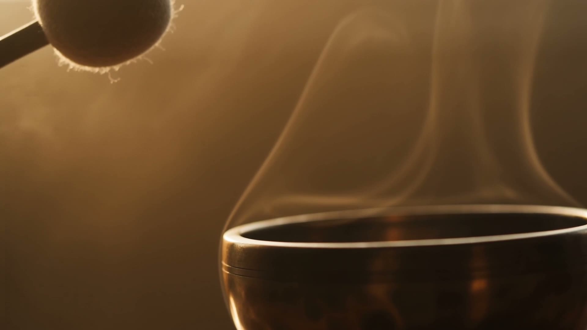An aged bronze singing bowl as a felt mallet approaches its rim in warm dawn light